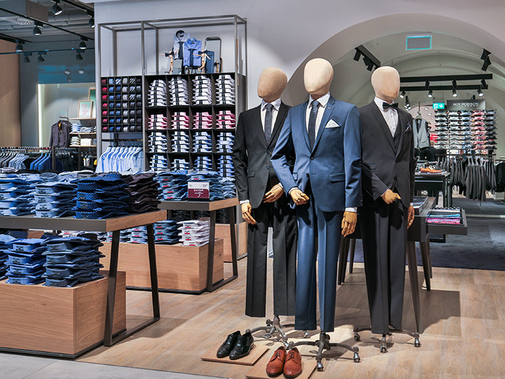 Ein Schaufenster in einem Bekleidungsgeschäft mit drei Schaufensterpuppen in formellen Anzügen. In der Umgebung sind ordentlich gestapelte Hemden und Accessoires zu sehen. Das Geschäft hat ein modernes Interieur mit sanfter Beleuchtung und gut sortierter Ware.