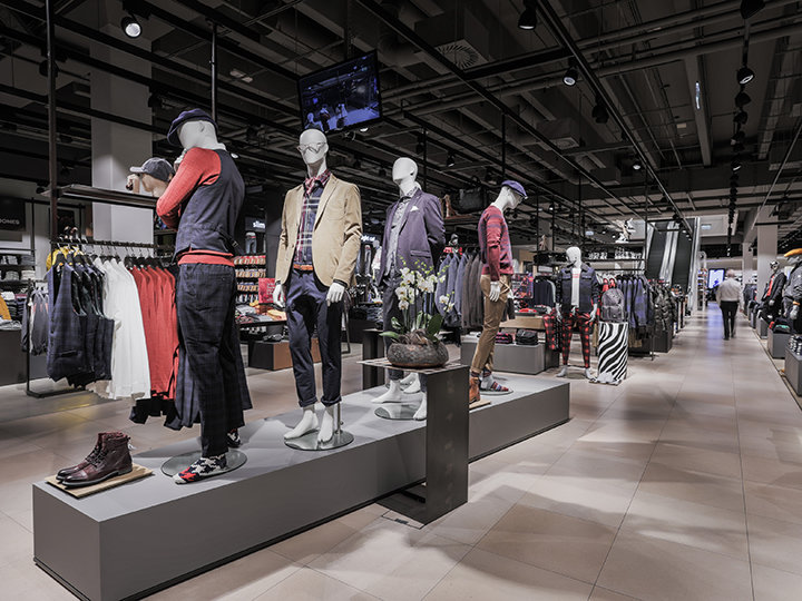 Ein Schaufenster in einem Bekleidungsgeschäft mit Schaufensterpuppen in verschiedenen Freizeit- und Abendgarderoben. Das Geschäft ist geräumig, im Hintergrund sind Kleiderständer mit Kleidung und Accessoires unter heller Beleuchtung. Eine Person geht durch den Gang.