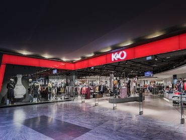 Schaufenster eines K&O-Modegeschäfts mit Schaufensterpuppen, die hinter großen Glasfenstern verschiedene Outfits präsentieren. Das Geschäft hat ein elegantes, modernes Design und ein leuchtend rotes Schild mit dem K&O-Logo, das prominent über dem Eingang angebracht ist.