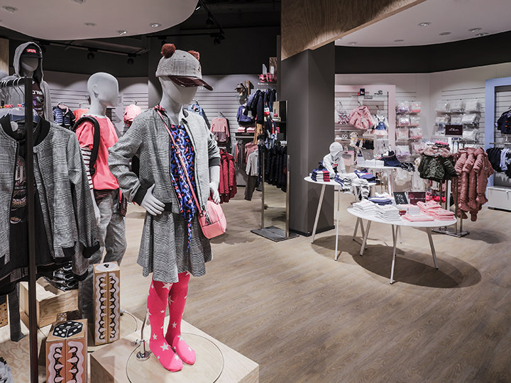 Ein Kinderbekleidungsgeschäft mit Schaufensterpuppen in farbenfrohen Outfits, darunter ein Hut und rosa Leggings. Auf Regalen und Ständern werden verschiedene Kleidungsstücke und Accessoires ausgestellt. Der Boden ist aus Holz und das Geschäft hat eine moderne Inneneinrichtung.