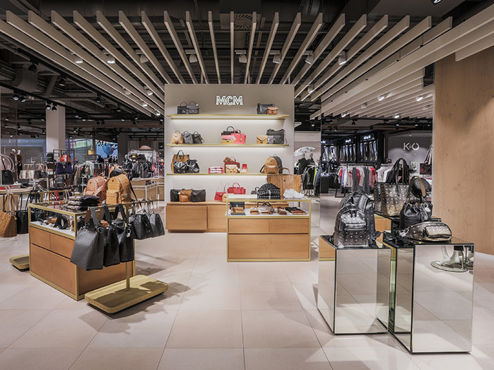 Ein modernes Ladeninneres, in dem eine Vielzahl von Luxushandtaschen auf Holz- und Glasdisplays präsentiert wird. Auf den Regalen an der Wand sind weitere Handtaschen zu sehen. Die Decke ist geometrisch gestaltet und die Beleuchtung ist hell, sodass die Waren hervorgehoben werden.