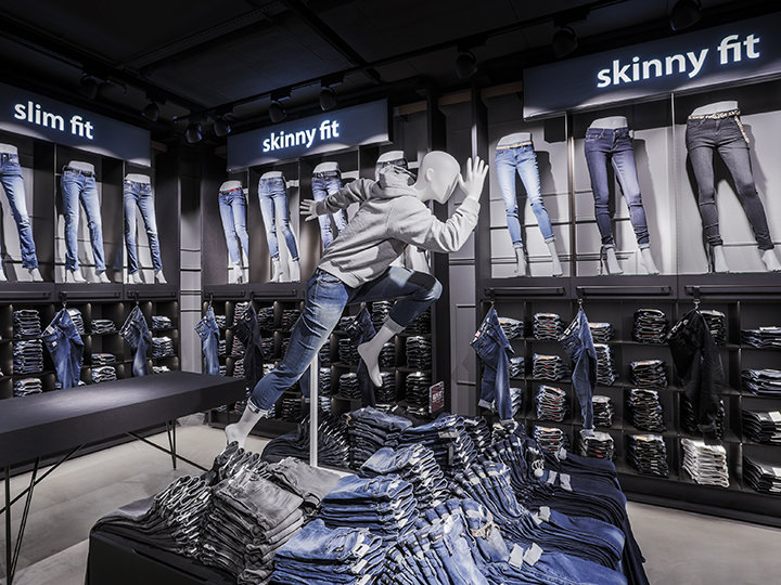 In einem Schaufenster werden verschiedene Jeans-Stile, darunter Slim Fit und Skinny Fit, präsentiert. Über einem Tisch steht eine Schaufensterpuppe in dynamischer Pose, sie trägt Jeans und einen Kapuzenpullover. Im Hintergrund sind die Regale mit gefalteten Jeans gefüllt.
