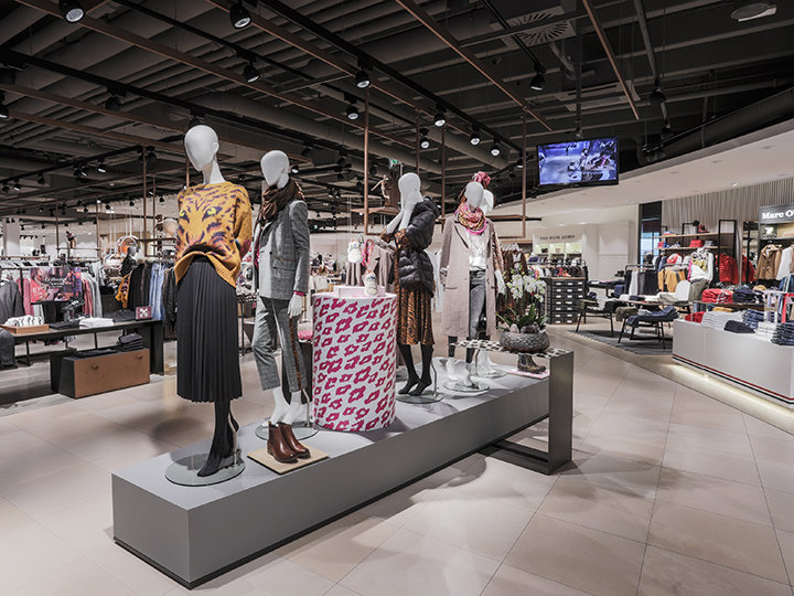 Ein stilvolles Interieur eines Bekleidungsgeschäfts mit Schaufensterpuppen in modischen Outfits. Das Geschäft ist modern eingerichtet, hat Kleiderständer und eine große gemusterte Kiste. Deckenbeleuchtung hebt verschiedene Displays hervor und im Hintergrund ist ein Bildschirm sichtbar.