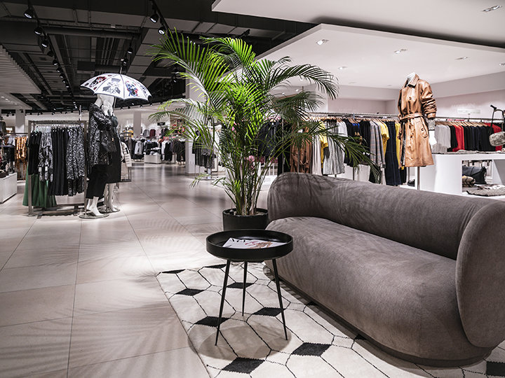 Ein modernes Interieur eines Bekleidungsgeschäfts mit Kleiderständern, darunter Mäntel und Jacken. Im Vordergrund stehen ein graues Sofa und ein runder schwarzer Tisch neben einer Topfpflanze. Im Hintergrund hält eine Schaufensterpuppe einen Regenschirm.