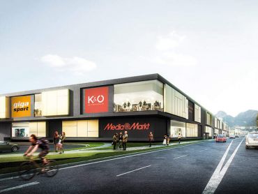 Ein modernes Einkaufszentrum mit verschiedenen Geschäften, darunter Media Markt. Menschen gehen spazieren und ein Radfahrer fährt auf der Straße vorbei. Das Gebäude ist von Gras umgeben und darüber ist ein klarer Himmel.