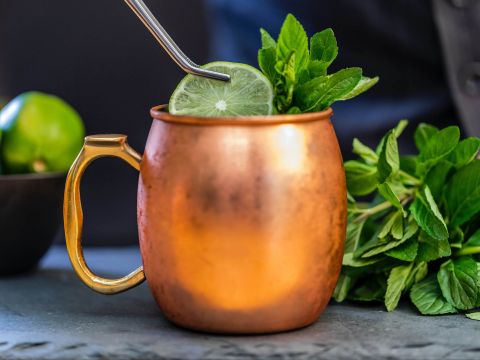 Ein Kupferbecher mit einem Getränk, garniert mit einer Limettenscheibe und frischen Minzblättern. In den Becher ist ein Metallstrohhalm gesteckt. Im Hintergrund sind eine Schale mit Limetten und ein Bund Minze zu sehen.