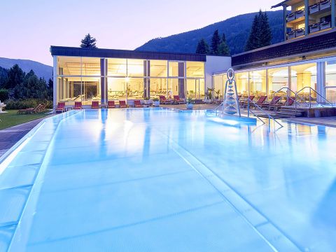 Ein moderner Außenpool mit leuchtend blauem Wasser in der Abenddämmerung. Der Pool ist von einer Terrasse mit Liegestühlen umgeben. Im Hintergrund ist ein großes Gebäude mit Glaswänden und eingeschalteten Lichtern zu sehen, vor einer Kulisse aus sanften Hügeln und Bäumen.