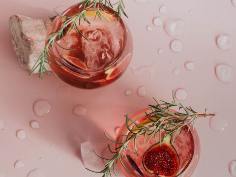 Zwei rosa Cocktails mit Eis, garniert mit Rosmarinzweigen und Feigenscheiben, stehen auf einer hellrosa Oberfläche. Ein Steinkeil stützt ein Glas. Wassertropfen verteilen sich und sorgen für eine erfrischende Note.