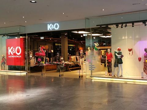 Schaufenster von Kastner & Öhler mit großen Glasfenstern, in denen bekleidete Schaufensterpuppen zu sehen sind. Im Ladeninneren sind Kleiderständer und eine Auswahl an Bekleidung zu sehen. Das K&O-Logo leuchtet deutlich sichtbar über dem Eingang.
