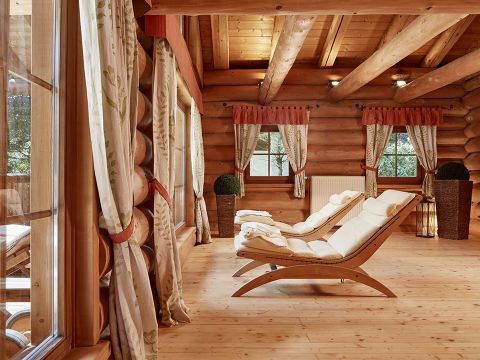 Ein gemütliches Blockhaus-Interieur mit großen Fenstern, hölzernen Deckenbalken und zwei gepolsterten Lounge-Sesseln auf einem polierten Holzboden. Vorhänge mit Blumenmuster rahmen die Fenster ein und im Raum sind dekorative Pflanzen verteilt.