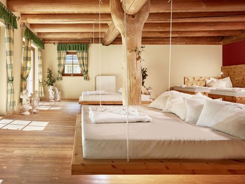 Ein gemütliches Zimmer mit Holzeinrichtung verfügt über drei Hängebetten mit weißer Bettwäsche. Sonnenlicht strömt durch große Fenster mit karierten Vorhängen und wirft Muster auf den Boden. Zwei kleine Statuen schmücken das Zimmer und verleihen ihm eine dekorative Note.