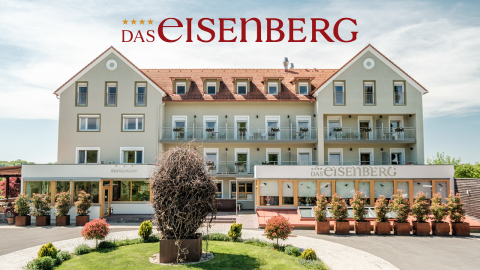 Ein großes cremefarbenes Gebäude mit rotem Dach und mehreren Balkonen vor einem klaren Himmel. Oben steht „Das Eisenberg“. Im Vordergrund ist ein Garten mit einer runden braunen Skulptur und Topfpflanzen in der Nähe des Eingangs zu sehen.