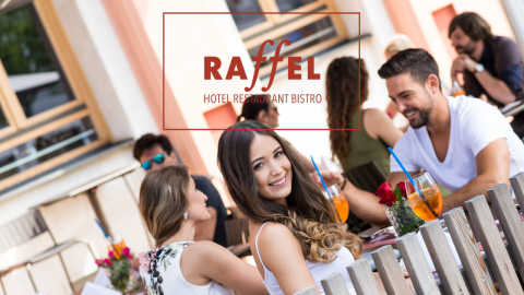 Eine Gruppe von Menschen sitzt draußen in einem Restaurant, genießt Getränke und unterhält sich. Eine Frau im Vordergrund lächelt in die Kamera. Auf dem Schild steht „Raffel Hotel Restaurant Bistro“.