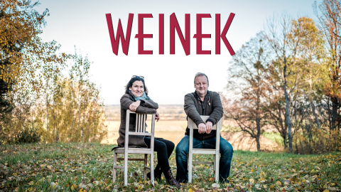 Ein Mann und eine Frau sitzen im Freien auf umgedrehten Holzstühlen und lächeln. Sie sind umgeben von herbstlichen Bäumen und einer grasbewachsenen Landschaft. Über ihnen prangt in großen roten Buchstaben das Wort „WEINEK“.