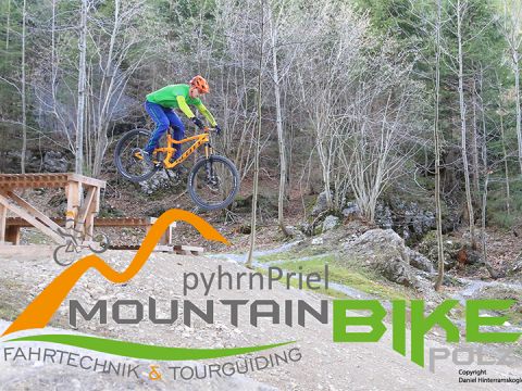Ein Mountainbiker in einer leuchtend grünen Jacke vollführt einen Sprung auf einem von Bäumen umgebenen Pfad. Die Worte „pyhrnPriel MOUNTAINBIKE“ und anderer Text überlagern das Bild und werben für Mountainbike-Fähigkeiten und -Touren.