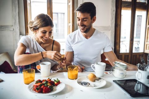 Ein Paar genießt sein Frühstück an einem Tisch in einem hellen Raum. Sie lächeln und haben eine Auswahl an Erdbeeren, Muffins und Orangensaft vor sich. Sonnenlicht fällt durch die großen Fenster herein und schafft eine warme und einladende Atmosphäre.