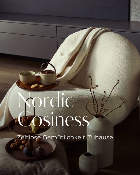 Ein gemütlicher Sessel mit einer cremefarbenen Decke, ein Tablett mit Tee und Snacks und eine minimalistische Einrichtung in einem modernen Wohnzimmer. Der Text lautet: "Nordische Gemütlichkeit. Zeitlose Gemütlichkeit Zuhause.".