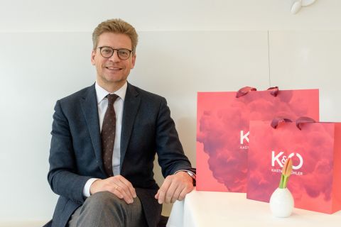 Ein Mann in Anzug und Brille sitzt lächelnd mit einer roten Einkaufstüte an einem Tisch. Auf dem Tisch steht auch eine kleine weiße Keramikvase.