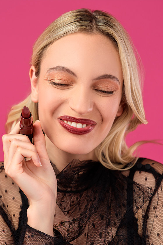 Eine Person mit blondem Haar lächelt und hält einen dunklen Lippenstift an die Wange. Sie trägt ein durchsichtiges schwarzes Oberteil mit Tupfenmuster vor einem hellrosa Hintergrund.