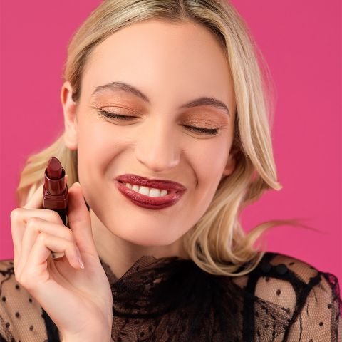 Eine Frau mit langen blonden Haaren lächelt mit geschlossenen Augen und hält einen dunkelroten Lippenstift an ihre Lippen. Sie trägt ein hauchdünnes schwarzes Spitzentop. Der Hintergrund ist rosa.