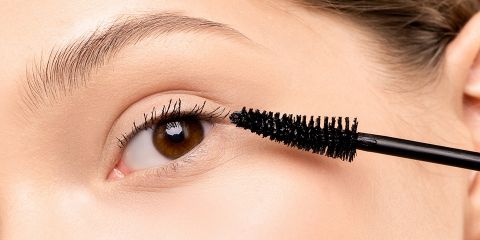 Eine Nahaufnahme einer Person, die schwarze Wimperntusche auf ihre oberen Wimpern aufträgt. Der Hintergrund hat eine neutrale Farbe und der Fokus liegt auf dem Auge und der Wimperntuschebürste.