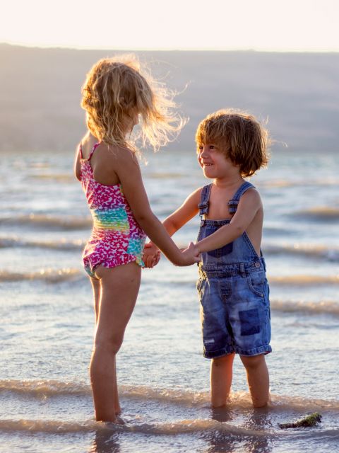 Zwei kleine Kinder halten Händchen und lächeln sich an, während sie im seichten Meerwasser stehen. Die Sonne scheint hinter ihnen und schafft eine warme und verspielte Atmosphäre. Eines trägt einen bunten Badeanzug, das andere einen Jeans-Overall.