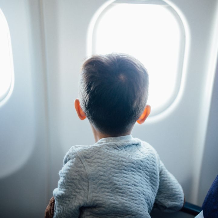 Ein kleines Kind in einem grauen Pullover blickt aus einem Flugzeugfenster. Das Innere des Flugzeugs ist sichtbar. Das Sonnenlicht fällt durch das Fenster und beleuchtet von hinten die kurzen Haare des Kindes.