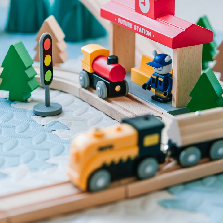 Eine hölzerne Spielzeugeisenbahn mit bunten Zügen auf Schienen. Eine Figur steht neben einem Gebäude mit rotem Dach und der Aufschrift „Future Station“. Auf der Spielmatte sind kleine Bäume, eine Ampel und ein Warnschild zu sehen.