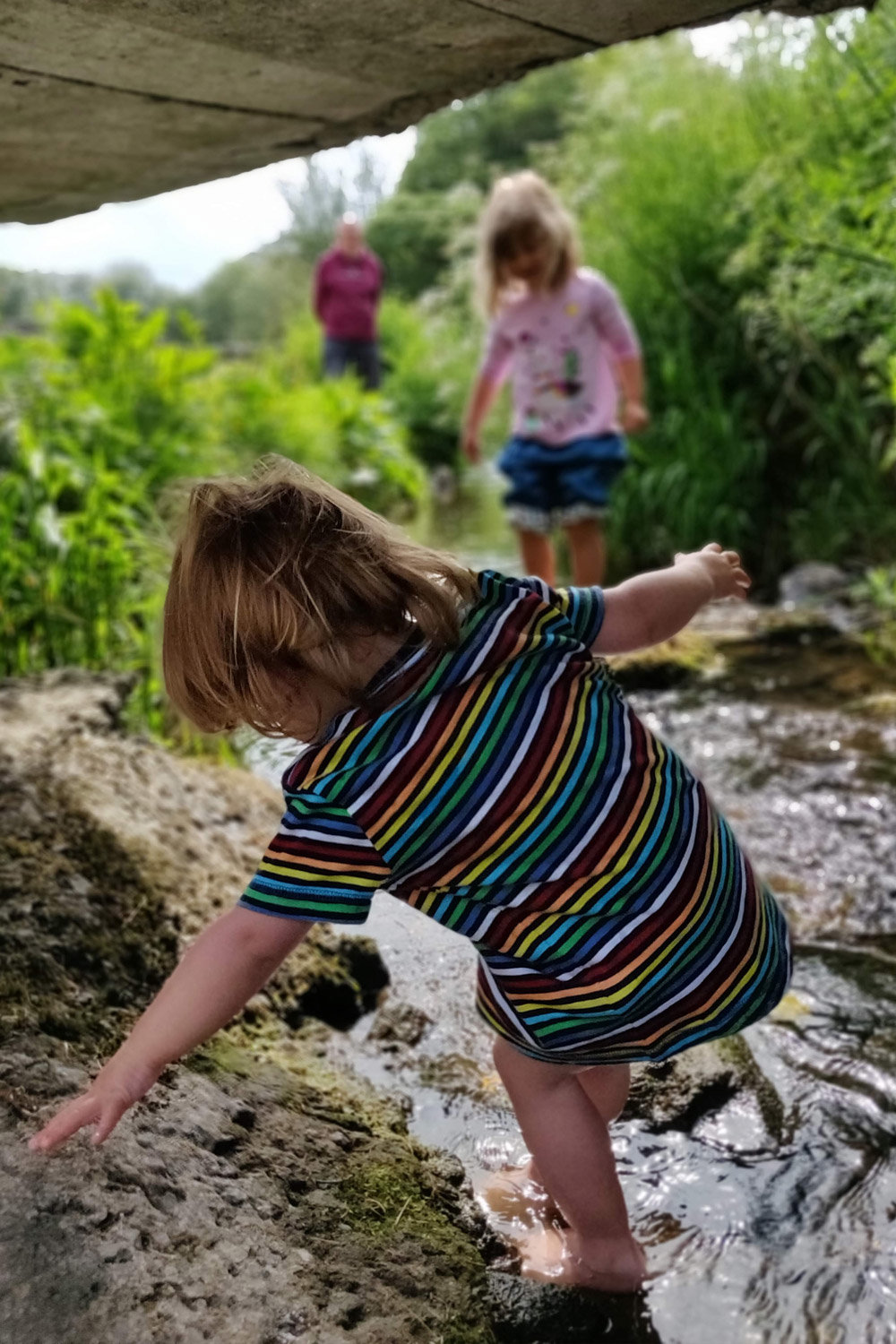 Zwei Kinder spielen in einem seichten Bach unter einer Brücke. Das Kind vorne trägt ein gestreiftes Hemd und steigt vorsichtig über das Wasser. Das Kind im Hintergrund trägt ein rosa Hemd. Sie sind von üppigem Grün umgeben und ein Erwachsener beobachtet sie aus der Ferne.