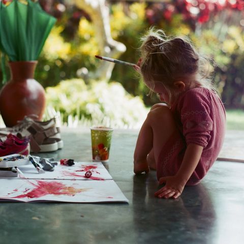 Ein kleines Kind sitzt auf dem Boden, umgeben von Malutensilien. Farben und Pinsel liegen verstreut auf Papier, leuchtende Farben sind zu sehen. Die Szene spielt im Freien, mit verschwommenem Grün und einem Tontopf im Hintergrund. Das Kind trägt ein rosa Outfit.
