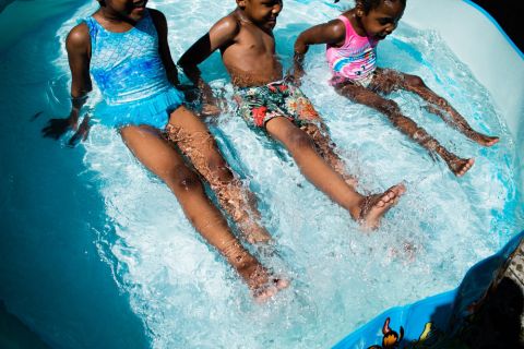 Drei Kinder spielen in einem kleinen Pool. Sie tragen bunte Badeanzüge. Sie sitzen mit ausgestreckten Beinen da, planschen im Wasser und genießen einen sonnigen Tag.