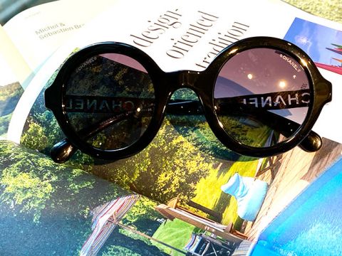 Runde schwarze Chanel-Sonnenbrillen liegen auf einem aufgeschlagenen Magazin mit Landschaftsaufnahmen. Die glänzend schwarzen Rahmen kontrastieren mit den farbenfrohen Natur- und Designbildern des Magazins.