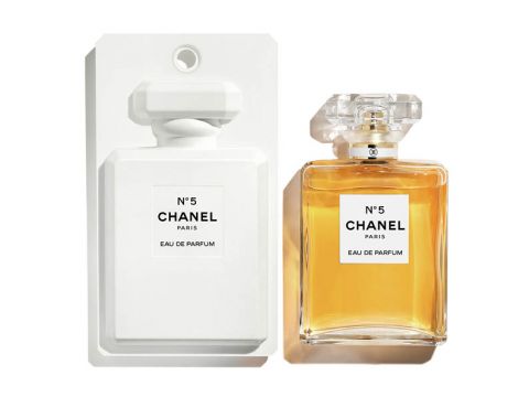 Eine Flasche Chanel No. 5 Eau de Parfum wird neben der weißen Verpackung ausgestellt. Das Parfüm befindet sich in einer klaren, rechteckigen Flasche mit facettiertem Verschluss und ist mit einer bernsteinfarbenen Flüssigkeit gefüllt. Das Etikett ist schlicht gehalten und zeigt das Chanel-Logo und den Produktnamen.