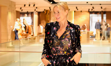 Eine Person mit blondem Haar steht lächelnd in einem hell erleuchteten Innenraum. Sie trägt ein Outfit mit Blumenmuster. Der Hintergrund ist leicht verschwommen und zeigt Kleidungsdisplays und andere Personen.
