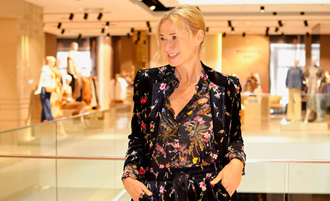 Eine Person mit blondem Haar steht lächelnd in einem hell erleuchteten Innenraum. Sie trägt ein Outfit mit Blumenmuster. Der Hintergrund ist leicht verschwommen und zeigt Kleidungsdisplays und andere Personen.