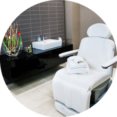 Ein moderner Spa-Raum mit einem weißen Liegesessel mit gefalteten Handtüchern, einer eleganten schwarzen Theke mit Waschbecken und einer dekorativen Vase mit roten Blumen vor einer grau gefliesten Wand.