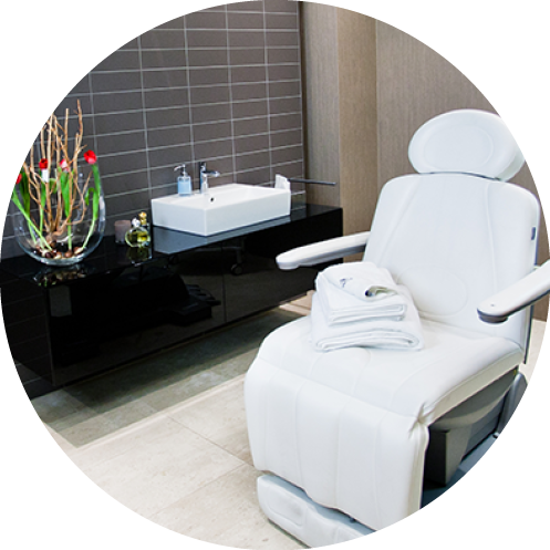 Ein moderner Spa-Raum mit einem weißen Liegesessel mit gefalteten Handtüchern, einer eleganten schwarzen Theke mit Waschbecken und einer dekorativen Vase mit roten Blumen vor einer grau gefliesten Wand.