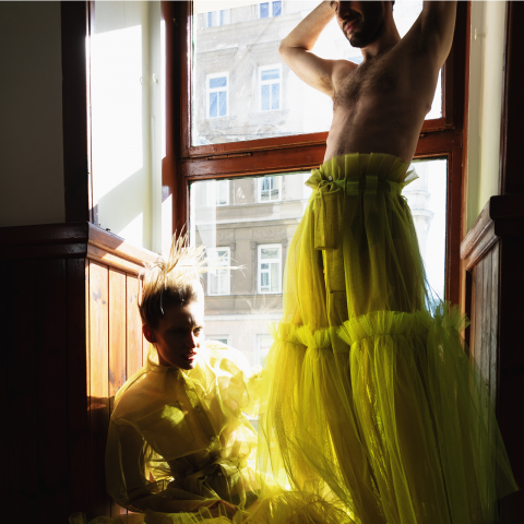 Eine Person in einem transparenten, leuchtend gelben Outfit posiert dramatisch an einem großen Fenster, das teilweise von Sonnenlicht erhellt wird. Eine andere Person steht in der Nähe und trägt ein ähnliches gelbes Tüllkleid. Von außen ist ein Gebäude zu sehen.