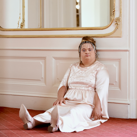 Eine Person mit langen Haaren und Kopfbedeckung sitzt auf dem Boden vor einer weiß getäfelten Wand. Sie trägt ein langes, elegantes weißes Kleid und rosa Schuhe. Über ihr hängt an der Wand ein großer Spiegel mit dekorativem Rahmen.