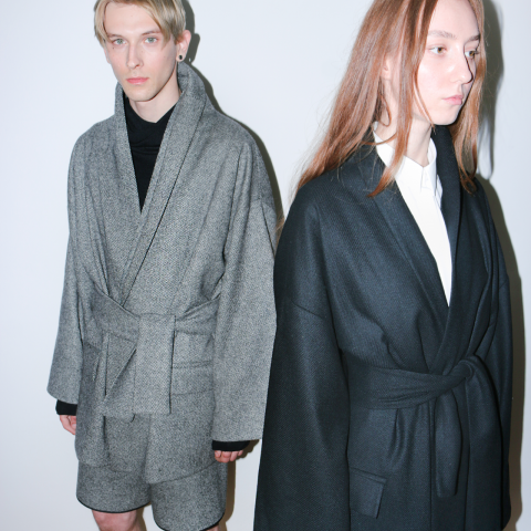 Zwei Models stehen vor einem weißen Hintergrund. Die Person links trägt eine graue Robe und Shorts, während die Person rechts eine schwarze Robe über einem weißen Hemd trägt. Beide haben glattes Haar und einen ernsten Gesichtsausdruck.