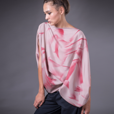 Eine Person trägt ein fließendes Oberteil mit rosa und roten Mustern und dazu dunkle Hosen. Sie steht vor einem grauen Hintergrund und blickt zur Seite. Das Oberteil hat ein asymmetrisches, drapiertes Design.