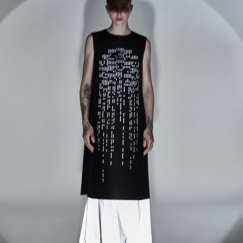 Eine Person steht in einem minimalistischen Studio und trägt ein ärmelloses schwarzes Kleid mit vertikalen, rechteckigen Ausschnittmustern und darunter einen weißen Rock. Die Beleuchtung erzeugt eine schattige Atmosphäre, die das Design des Kleides und die Tattoos des Models hervorhebt.
