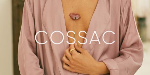 Eine Person in einem seidigen, hellrosa Gewand hält einen rosa Blütenstiel zwischen den Fingern in der Nähe ihrer Brust. Das Wort „COSSAC“ ist in weißer Schrift über das Bild gelegt.
