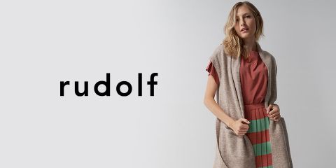 Eine Frau, die eine ärmellose Strickjacke über einem rostroten Hemd und einem gestreiften Rock trägt, steht selbstbewusst vor einem schlichten Hintergrund. Neben ihr steht in fetten Kleinbuchstaben das Wort „Rudolf“.