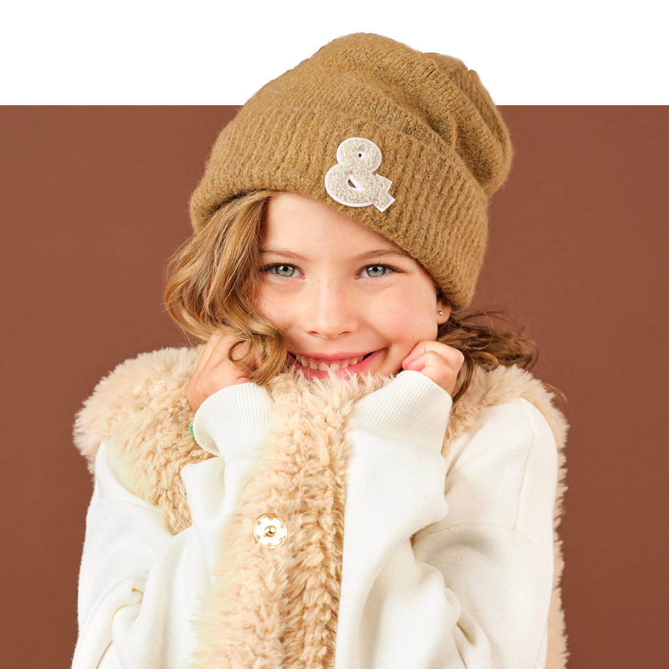 Ein lächelndes Kind trägt eine braune Strickmütze mit einem Et-Zeichen-Aufnäher. Es hat ein weißes Oberteil und eine flauschige beige Weste und steht vor einem braunen Hintergrund.