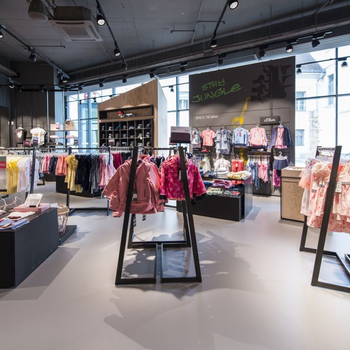 Ein hell erleuchtetes Kinderbekleidungsgeschäft mit Regalen voller farbenfroher Outfits. Verschiedene Oberteile, Kleider und Jacken sind ordentlich angeordnet und darüber hängen Schilder mit verspielten Motiven. Große Fenster lassen Tageslicht herein und schaffen eine einladende Atmosphäre.