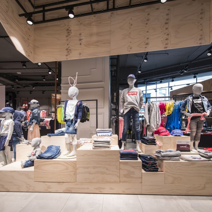 Ein modernes Schaufenster für ein Bekleidungsgeschäft mit Schaufensterpuppen in legerer Kleidung. Die Schaufensterpuppen stehen auf einer abgestuften Holzplattform, umgeben von Kleiderständern und Accessoires. Das Geschäft ist hell und geräumig und hat ein minimalistisches Design.