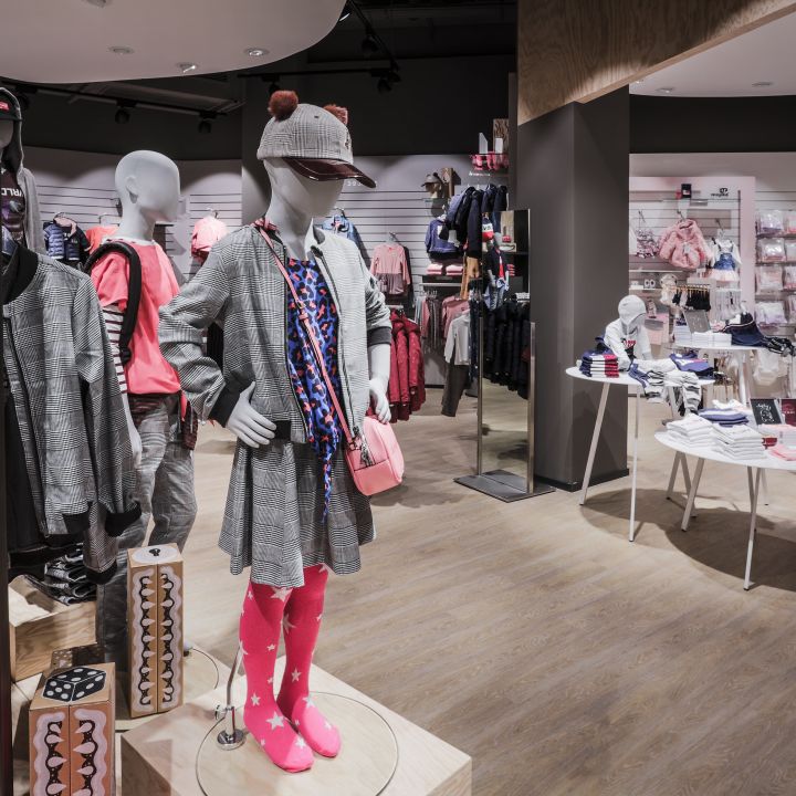 In einem Kinderbekleidungsgeschäft werden auf Schaufensterpuppen Outfits ausgestellt. Die Schaufensterpuppe im Vordergrund trägt einen karierten Mantel, rosa Stiefel und eine Mütze. Auf Regalen und Tischen sind ordentlich Kleidungsstücke in verschiedenen Farben und Stilen angeordnet. Der Laden hat Holzböden und eine warme Beleuchtung.