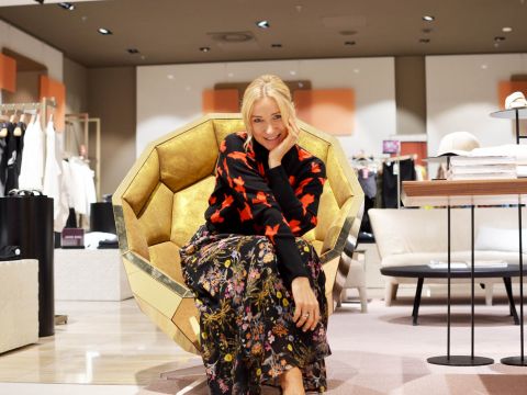 Eine Frau sitzt auf einem modernen, goldenen, geometrischen Stuhl in einem stilvollen Geschäft. Sie trägt ein schwarzes Oberteil mit roten Mustern und einen bunten Blumenrock und lächelt in die Kamera. Das Geschäft ist mit Kleiderständern, Regalen und zeitgenössischen Möbeln ausgestattet.
