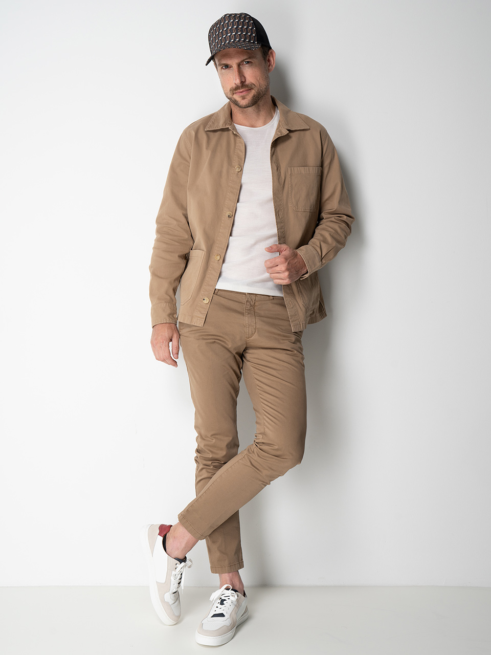 Ein Mann in beiger Jacke, passender Hose, weißem Hemd und schwarzer Mütze posiert vor einer weißen Wand. Seine Hände stecken in den Taschen und er trägt weiße Turnschuhe.